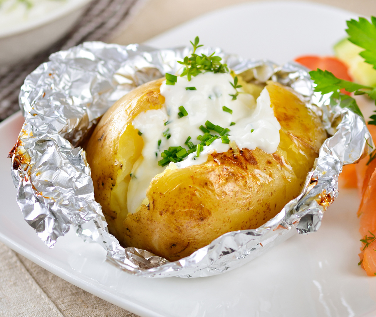 Запеченный картофель в фольге со сметаной и рубленым зеленым луком, подается на белой тарелке с гарниром из овощей и зелени от Soldis.