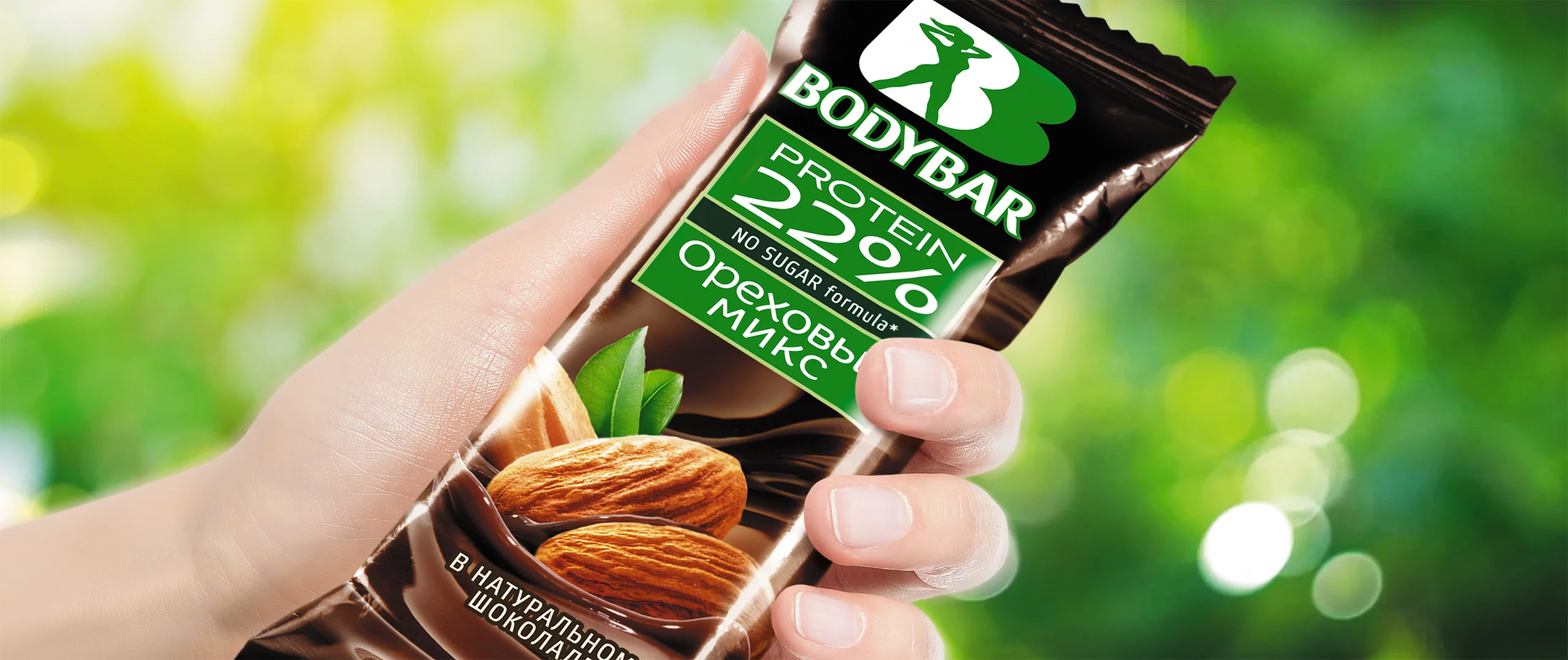 Рука держит протеиновый батончик BODYBAR с 22% белка от Soldis. На упаковке изображены миндаль, зеленые листья и текст на русском и английском языках. Размытая зелень и солнечный свет заполняют фон.