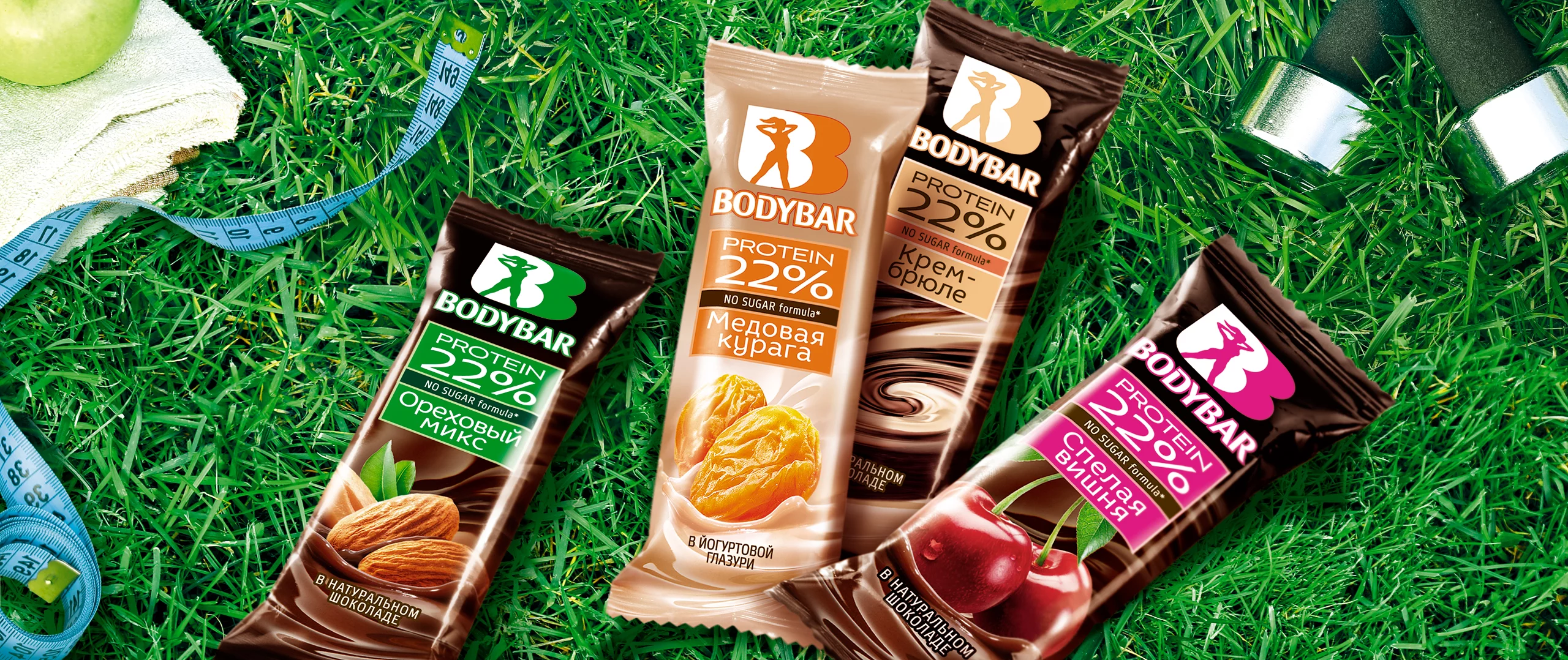 Четыре протеиновых батончика BODYBAR (со вкусами миндаля, абрикоса, шоколада и вишни) от Soldis выставлены на траве на фоне теннисного мяча, синей рулетки и черной ручки.