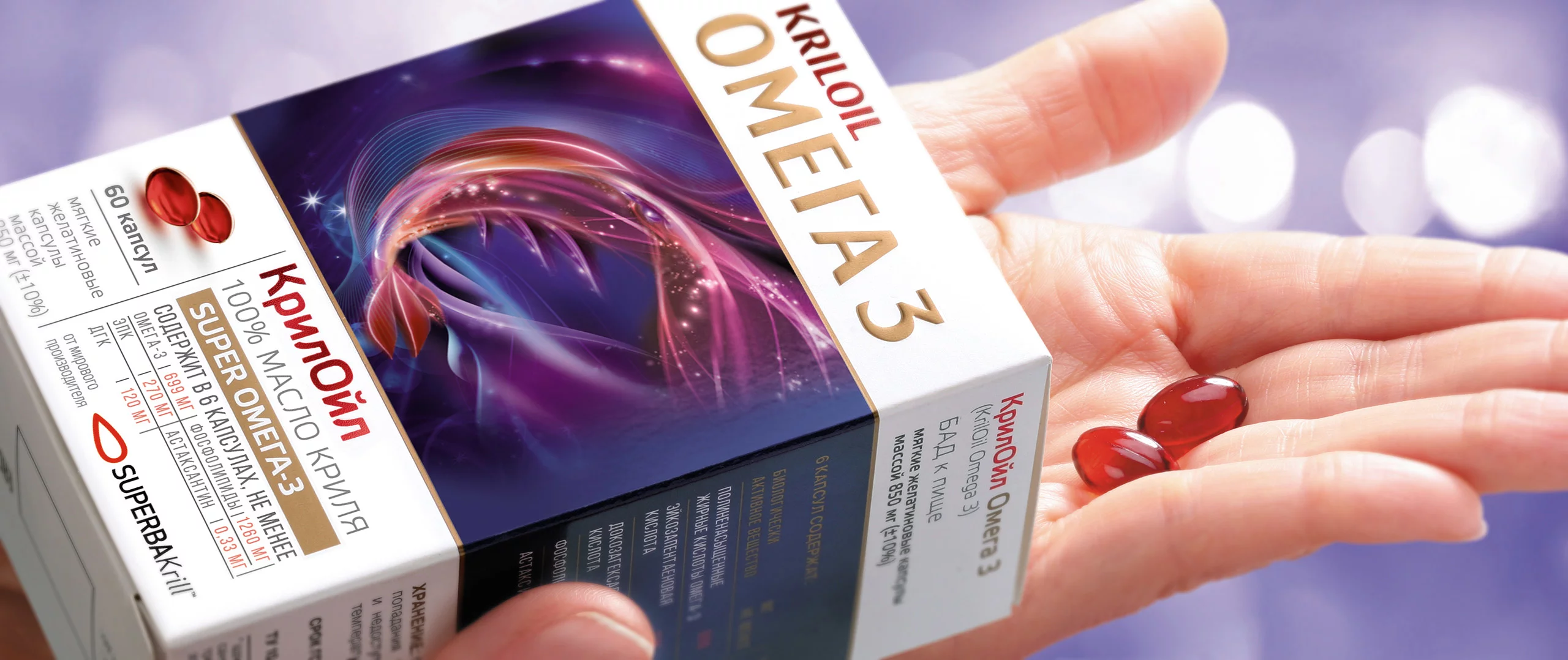 Рука держит три красные мягкие капсулы рядом с коробкой с надписью KRILOIL OMEGA 3 на русском и английском языках, на упаковке изображена красочная абстрактная графика Солдиса.