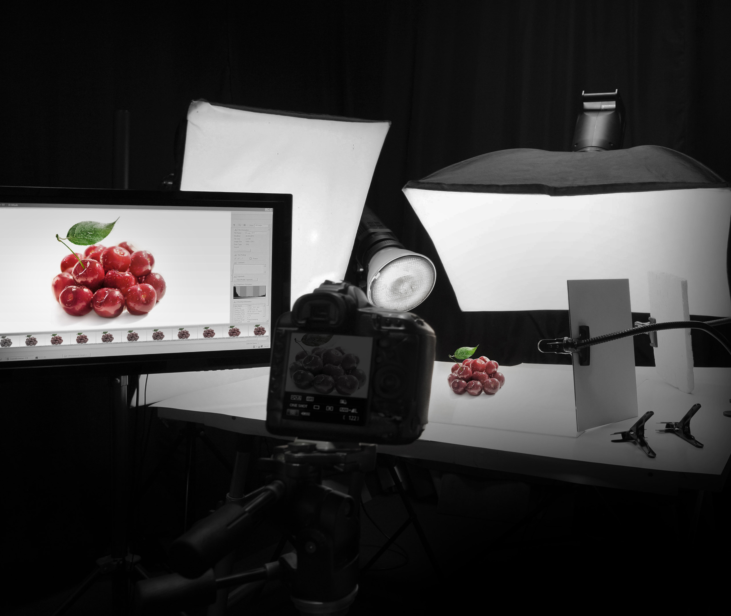 Профессиональная фотоустановка Soldis снимает красные вишни на белом столе, окруженном студийным освещением, отражателями и камерой, а изображение выводится на монитор компьютера.