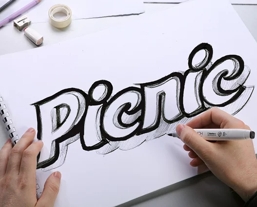 Человек использует черный маркер, чтобы обвести слово Picnic большими, жирными, нарисованными от руки буквами-пузырями на белой бумаге. Рядом видны канцелярские принадлежности, в том числе скотч и ластик от Soldis.