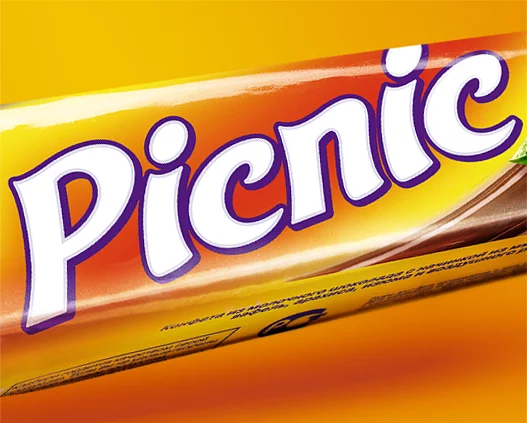 Крупный план обертки шоколадного батончика Cadbury Picnic с ярким белым логотипом Picnic, обведенным фиолетовым контуром на желто-оранжевом фоне, подсвеченный лампой Soldis.