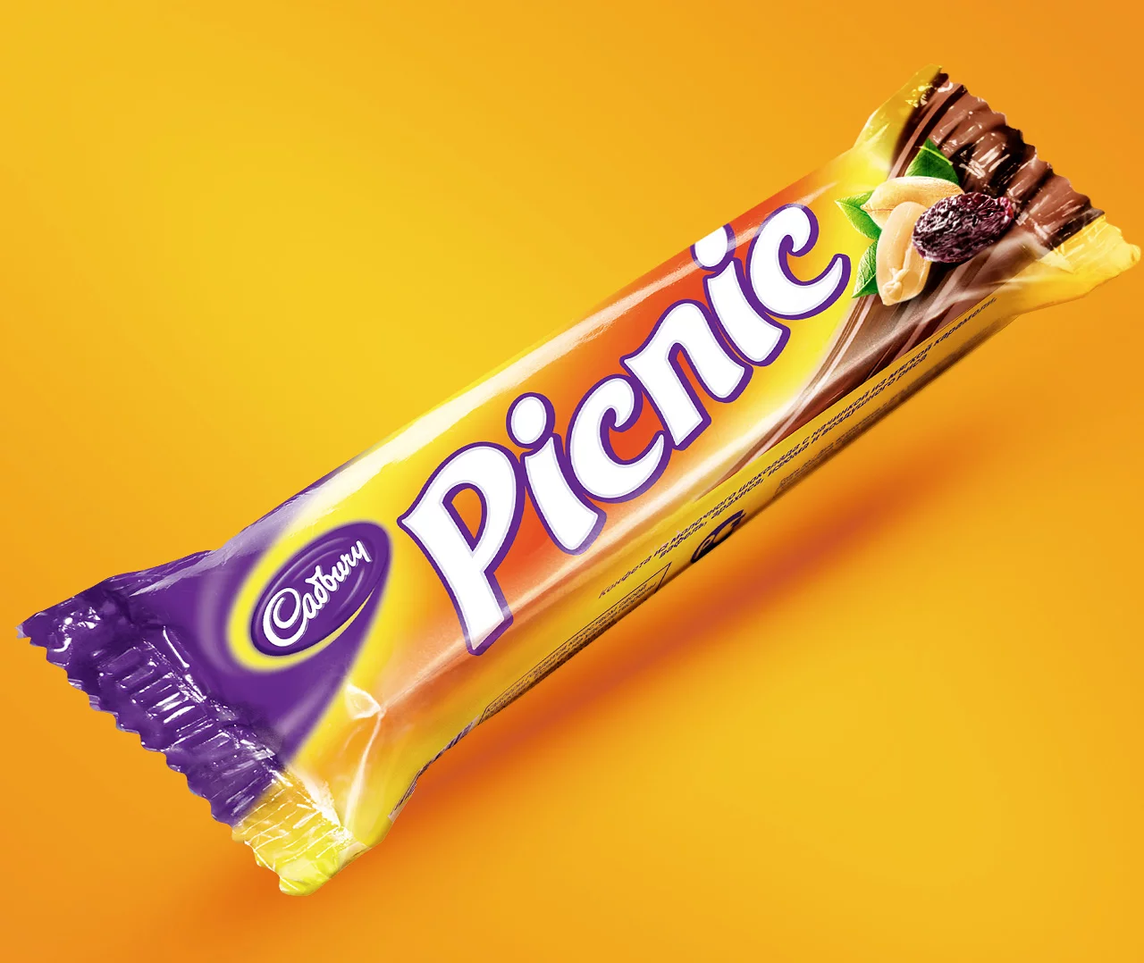 Шоколадный батончик Cadbury Picnic в желто-фиолетовой упаковке изображен на ярко-оранжевом фоне. Обертка, разработанная Soldis, отображает название бренда вместе с изображениями арахиса и изюма.