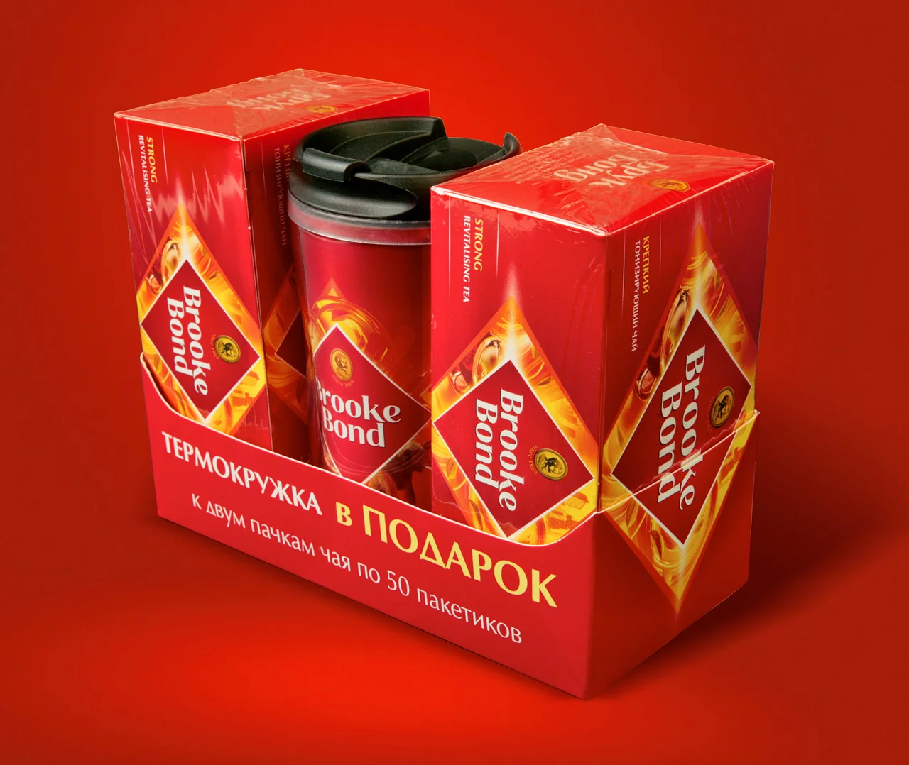 Подарочный набор чая Brooke Bond от Soldis включает две красные коробки чая и кружку для путешествий в центре, все упаковано вместе на красном фоне. Русский текст на упаковке упоминает кружку-термос в качестве подарка.