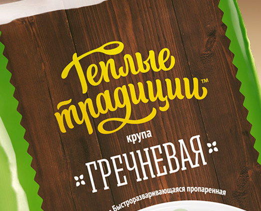 На зеленой упаковке продуктов питания на деревянном фактурном фоне изображен желтый стилизованный кириллический текст с надписью «Теплые традиции» и белый текст с надписью «Крупа Гречневая», обозначающий гречневую крупу «Солдис».