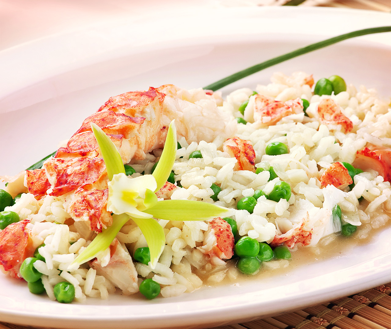 Тарелка риса, смешанного с зеленым горошком и кусочками лобстера, украшенная желтым цветком и длинным зеленым луком, элегантно подана на белом блюде от Soldis.