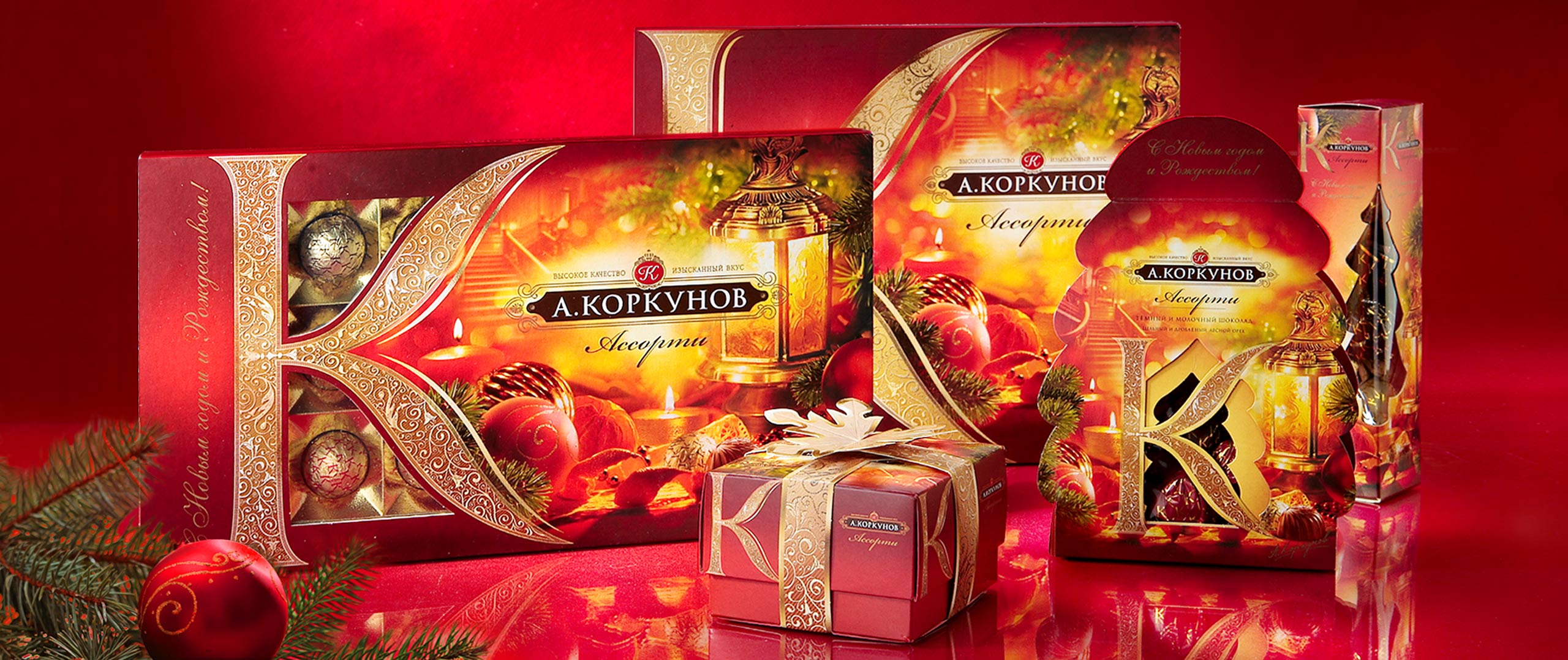 Ассорти из подарочных коробок с шоколадом «А. Коркунов» от Soldis, украшенных золотыми узорами и праздничными орнаментами на красном фоне, с сосновой веткой и рождественской елкой на переднем плане.