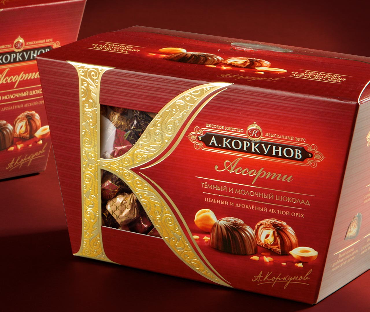 Красно-золотая коробка шоколадных конфет «А. Коркунов» от Soldis с большой декоративной буквой «К» на лицевой стороне, изображениями различных шоколадных изделий и окошком, демонстрирующим шоколадные конфеты внутри.