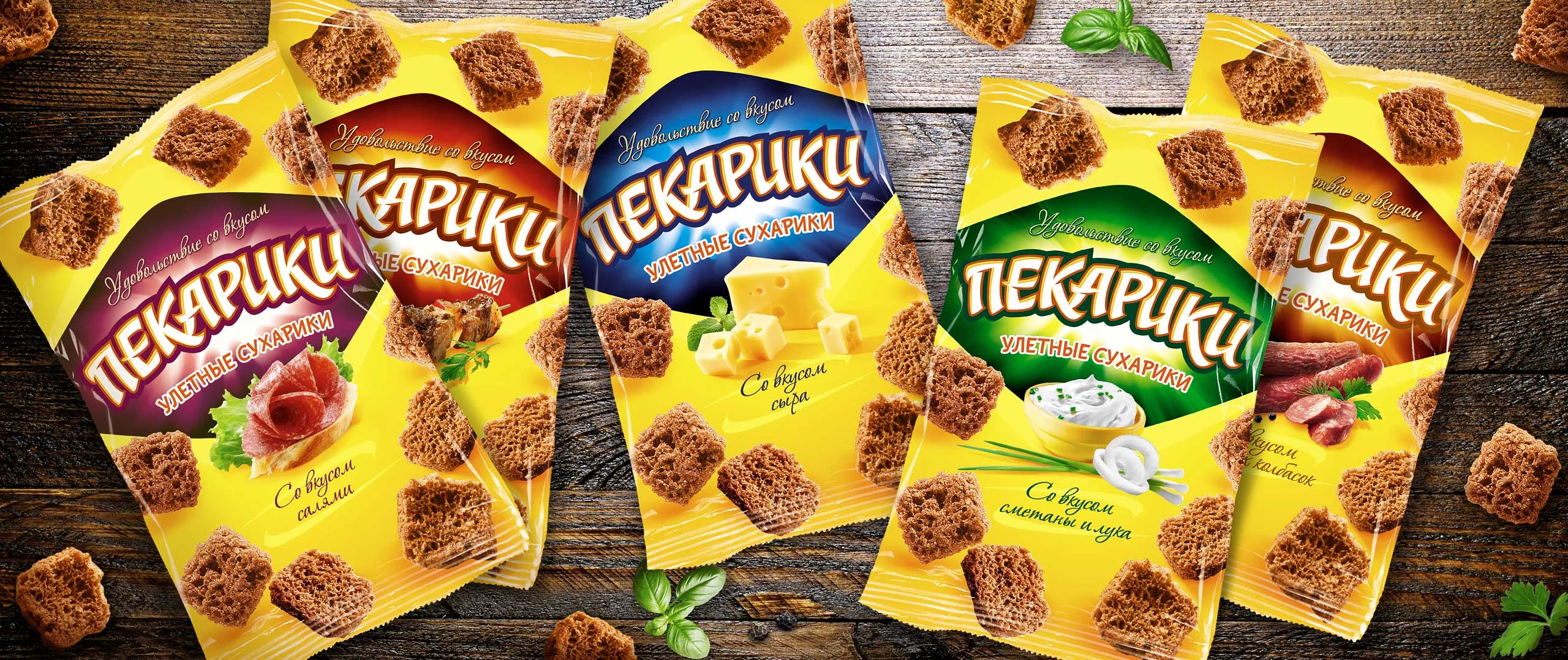 Четыре желтые упаковки русских ржаных сухариков «Soldis» со вкусом салями, сыра, зелени и острой колбасы выставлены на деревянной поверхности, а вокруг разбросаны отдельные сухарики.