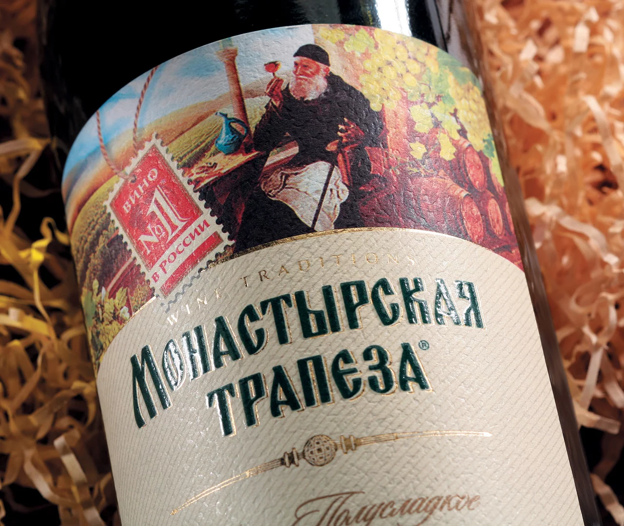 Крупный план бутылки вина в дизайне от Soldis с этикеткой «Монастырская Трапеза», на которой изображен бородатый мужчина в черной мантии, сидящий среди виноградников в окружении упаковочного материала, похожего на солому.